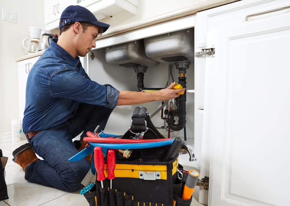 Plumber repairing drain and supply pipes beneath a double kitchen sink